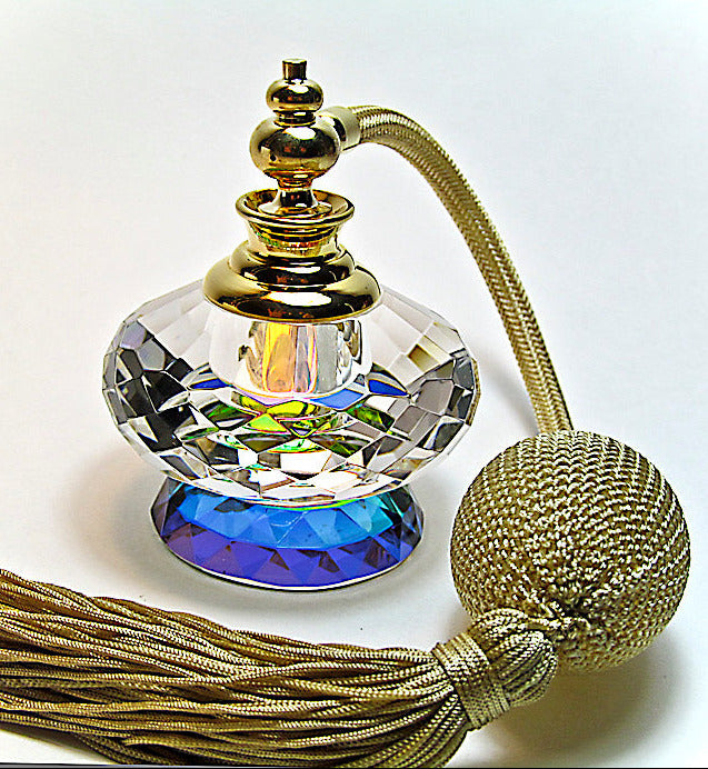 A decorative perfume bottle with a faceted clear crystal body, a blue iridescent base, a gold-toned cap and neck, a gold braided neck cord, and a large gold tassel on a white background.