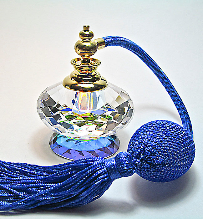 A faceted clear glass perfume bottle with a gold-toned cap and a long blue tassel with a knotted ball, photographed on a plain light background.