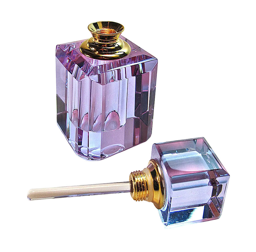 A faceted purple glass perfume bottle with a gold-toned cap, shown next to its detached glass dropper pipette on a light beige background.